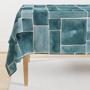 Puede incluir: Un mantel de mesa azul verdoso con un patrón de azulejos rectangulares en acuarela. El mantel está drapeado sobre una mesa de madera.