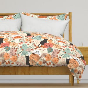 Può includere: Una federa per piumone di colore crema con una stampa a tema Halloween che presenta gatti neri, zucche, fantasmi e fiori in tonalità arancio, rosa e verde.