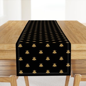 May include: Black table runner with a repeating pattern of gold-colored bees. The runner is rectangular and has a white border. The runner is displayed on a wooden table.