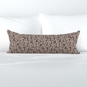 May include: A long, rectangular decorative pillow with a floral pattern. The pillow features a dark brown background with a repeating pattern of light beige flowers and leaves. The pillow is placed on a white bed.