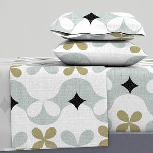 May include: Two decorative pillows and a folded sheet with a repeating pattern. The pattern features light blue, olive green, and black diamond shapes, and floral designs on a white background. The pillows are stacked on top of the sheet, creating a layered effect.