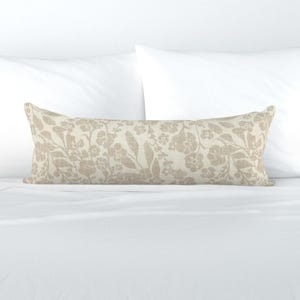 May include: A long, rectangular lumbar pillow featuring a light beige fabric base with a repeating pattern of tan floral and leaf motifs. The pillow has a textured appearance and is positioned horizontally against a plain white background.