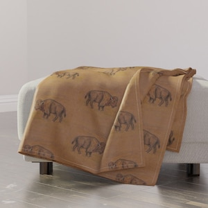 May include: A brown throw blanket with a repeating pattern of black buffalo silhouettes. The blanket is folded and draped over a white couch.