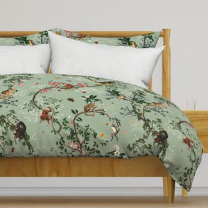 May include: A light green duvet cover and pillowcases featuring a whimsical design of monkeys in a tree. The bedding set has a botanical theme with branches, flowers, and butterflies. The bed frame is made of light wood.