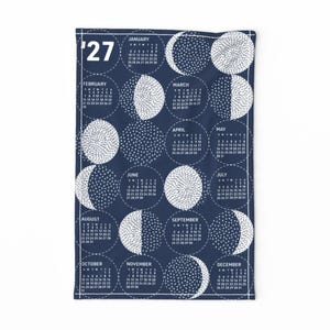 May include: A navy blue tea towel featuring a white 2027 calendar design. The layout includes twelve circular sections, each representing a month with its corresponding dates and a unique white graphic pattern depicting various moon phases.
