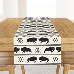 May include: A black and white table runner with a repeating pattern of buffalo silhouettes and geometric designs. The runner is on a wooden table.