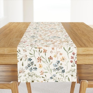 Puede incluir: Un camino de mesa floral con fondo crema y un estampado de flores silvestres coloridas en tonos rosa, azul, naranja y verde. El camino de mesa se muestra sobre una mesa de madera.
