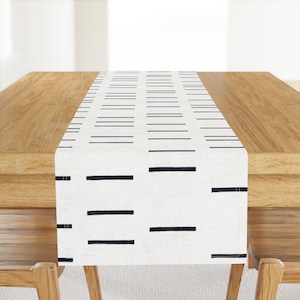 May include: White table runner with a black and white geometric pattern of horizontal lines. The runner is on a wooden table.