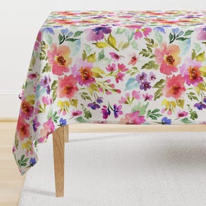 May include: A white tablecloth with a watercolor floral pattern in shades of pink, purple, and green. The tablecloth is draped over a table with wooden legs.