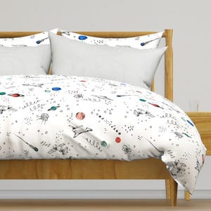 Op de afbeelding: Wit dekbedovertrek met een ruimtethema met een patroon van planeten, sterren, raketten en kometen in zwart, blauw, groen en rood.