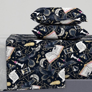 Op de afbeelding: Een zwart en gouden beddengoedset met een herhalend patroon van raven, sterren, manen, sleutels en open boeken. De beddengoedset bevat een plat laken, een hoeslaken en twee kussenslopen.