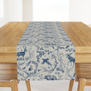 May include: A blue and white table runner with a woodland toile pattern. The design features deer, birds, rabbits, and bears among branches and foliage. The runner is displayed on a wooden table.