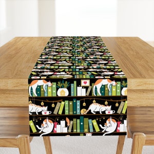 May include: A black table runner with a repeating pattern of cats sleeping on bookshelves. The bookshelves are filled with colorful books and there are small potted plants and candles on the shelves. The cats are white with orange markings.