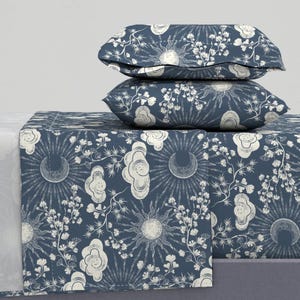 Op de afbeelding: Een marineblauwe beddenset met een herhalend patroon van zonnestralen, wolken en bloemelementen in wit. De set bevat twee kussens die op een bijpassend sprei zijn gestapeld. Het ontwerp creëert een samenhangende en decoratieve esthetiek.
