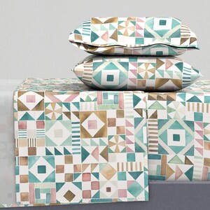 May include: Two decorative pillows and a bed runner with a geometric pattern in teal, pink, brown, and gold. The pattern features squares, triangles, and other shapes on a white background. The pillows are stacked on the bed runner.