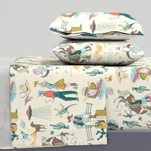 May include: Cream-colored bedding set with a repeating pattern of cowboys, cacti, horses, and UFOs in shades of blue, red, and brown. The set includes two pillows and a fitted sheet, creating a whimsical, Western-themed bedroom decor.