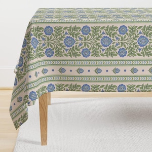 May include: A beige tablecloth with a repeating floral pattern in blue and green. The design features stylized blue flowers and green leaves, with a border of arrows and dots. The tablecloth is draped over a wooden table.