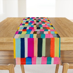 May include: A colorful table runner with a geometric pattern of vertical stripes in various shades of pink, blue, green, yellow, orange, and brown. The runner is made of fabric and has a white border.
