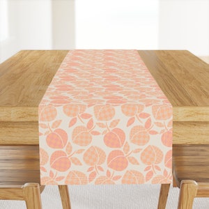 May include: A peach-colored table runner with a repeating pattern of peach fruit and leaves. The runner is on a wooden table.