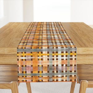 May include: A rectangular table runner with a woven pattern in shades of orange, brown, gray, and yellow. The runner is placed on a wooden table, adding a decorative touch to the dining area.