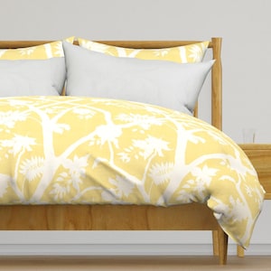 May include: A bed with a light wood frame and white pillows. The bedding features a yellow and white tree pattern. The duvet cover and pillowcases display a repeating design of stylized trees and foliage.