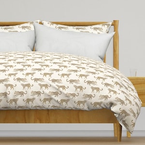 May include: A duvet cover and matching pillow shams featuring an all-over repeating pattern of brown and tan leopards on a cream-colored background. The bedding set is draped over a wooden bed frame with two solid white rectangular pillows.