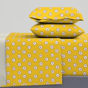 Puede incluir: Una sábana de cama amarilla con un patrón de margaritas blancas y dos fundas de almohada a juego. La sábana y las fundas de almohada están hechas de tela suave y cómoda.