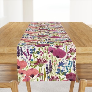 May include: A floral table runner featuring a watercolor design of pink, purple, and blue flowers on a cream background. The runner is placed on a wooden table, showcasing the vibrant botanical pattern. The runner's dimensions are suitable for a dining table.