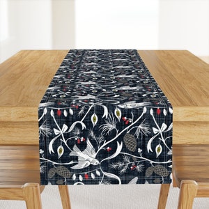 May include: A navy blue table runner with a white and gray winter-themed pattern. The pattern includes birds, pine branches, pine cones, berries, and ornaments.