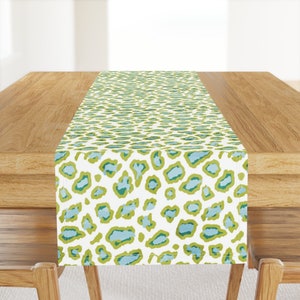 Puede incluir: Un camino de mesa blanco con un estampado de leopardo verde y azul. El camino de mesa está sobre una mesa de madera.