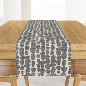 May include: A white table runner with a gray abstract pattern of small circles and ovals. The runner is on a wooden table.