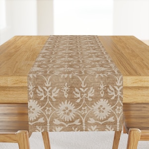 May include: A beige table runner with a white floral pattern. The runner is on a wooden table.