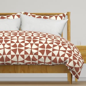 May include: A bed with a wooden frame and a duvet cover featuring a geometric pattern in terracotta and cream. Two white pillows and a matching pillowcase are visible. The design is a repeating pattern of semi-circles.