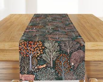 May include: A rectangular table runner featuring a dark forest pattern with a black background. The design includes various trees with orange, green, and white leaves, interspersed with illustrations of a brown bear, a fox, and a deer.