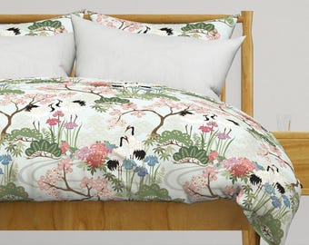 Op de afbeelding: Een dekbedovertrek en bijpassende kussenslopen met een lichtblauwe achtergrond en een herhalend patroon van witte kraanvogels met zwarte accenten, roze kersenbloesems, groene dennenbomen en rode en blauwe bloemen op een houten bedframe.