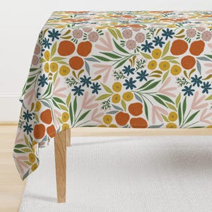 May include: A white tablecloth with a floral pattern featuring orange, yellow, green, blue, and pink flowers. The tablecloth is draped over a table with wooden legs.