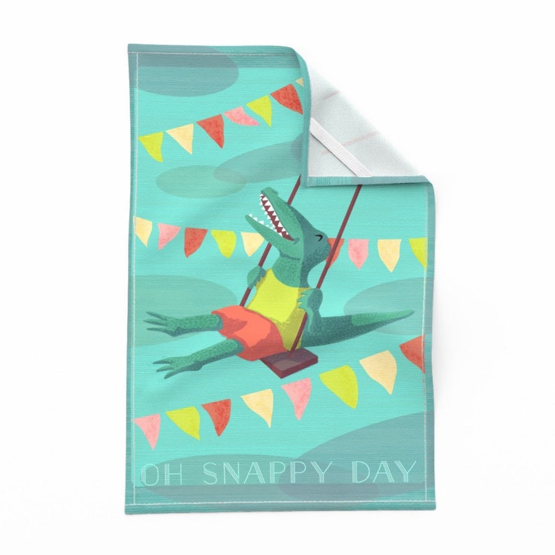 May include: A teal blue kitchen towel with a green alligator on a swing. The alligator is wearing a yellow shirt and red shorts. The background is decorated with colorful flags and the text "OH SNAPPY DAY".