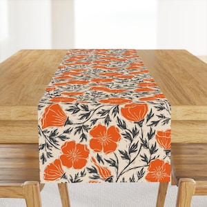 May include: A rectangular table runner featuring a repeating floral pattern. The design showcases large orange poppy flowers with black stems and leaves against a cream background. The runner is displayed on a wooden table.
