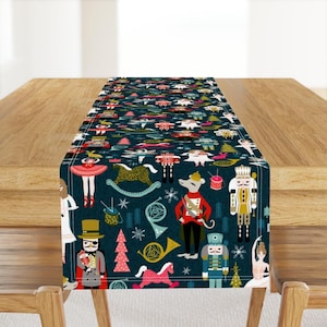 May include: A dark blue table runner featuring a repeating pattern of nutcrackers, ballerinas, rocking horses, musical instruments, and Christmas trees in red, gold, white, and teal. The design includes stylized figures and festive holiday motifs.