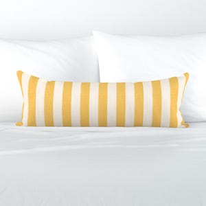 May include: A rectangular decorative pillow with vertical stripes in yellow and off-white. The pillow is placed on a white bed with white pillows in the background. The striped pattern adds a touch of color and design to the bedding.
