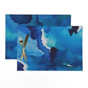 May include: Two blue watercolor placemats with abstract designs. The placemats are made of fabric and have a stitched edge.