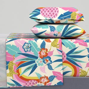May include: Bedding set featuring a vibrant tropical pattern. The design includes pink pineapples, blue leaves, and orange flowers on a cream background. Two pillows are stacked on top of the folded sheet set, showcasing the colorful print.