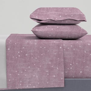 May include: A dusty rose bedding set featuring a fitted sheet, flat sheet, and two pillowcases. The fabric is patterned with small white stars. The pillowcases are stacked and have a decorative stitched edge. The sheets are made of a soft, textured material.
