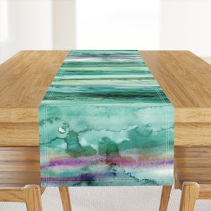 May include: A teal and green watercolor table runner with a striped pattern. The runner has a subtle purple and pink stripe at the bottom.