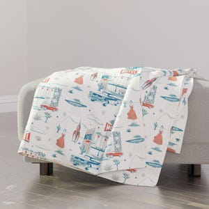 May include: White throw blanket with a retro design featuring illustrations of motels, cars, rockets, UFOs, and women in dresses. The design is in shades of blue, red, and gray.