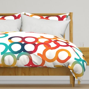 May include: A white duvet cover and matching pillow shams featuring a repeating pattern of interlocking circles in vibrant shades of red, orange, yellow, and teal. The bedding is displayed on a light brown wooden bed frame with a headboard.
