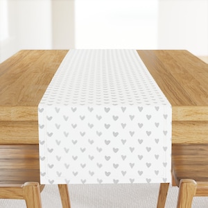 May include: White table runner with a repeating pattern of gray hearts. The runner is on a wooden table.