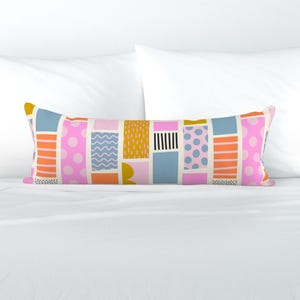 May include: A long rectangular cushion with a geometric pattern. The pattern is made up of various coloured rectangles, including pink, orange, yellow, blue, and white. The rectangles have different patterns, such as polka dots, stripes, and wavy lines.