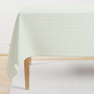 May include: A rectangular tablecloth featuring a light sage green and white gingham check pattern. The fabric drapes over the edges of a wooden table, displaying a consistent grid design across the entire surface of the textile covering.