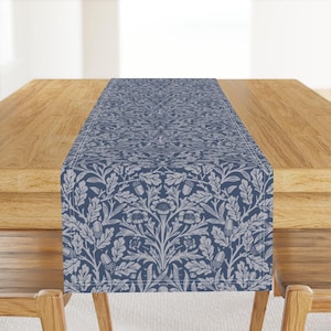 Può includere: Runner da tavolo floreale blu e bianco con un motivo ripetuto di foglie, ghiande e fiori. Il runner è esposto su un tavolo di legno, evidenziando il suo design e la sua consistenza. Il runner è largo circa 30 cm.